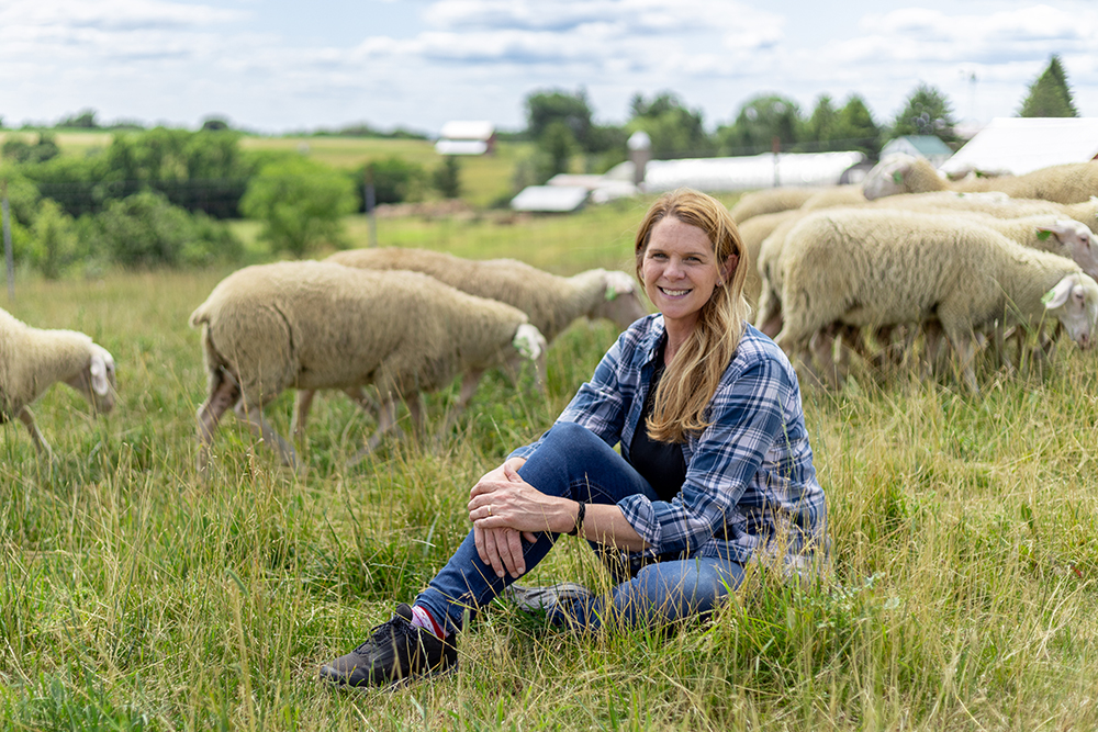 A woman sitting in a grassy farm field surrounded by grazing sheep