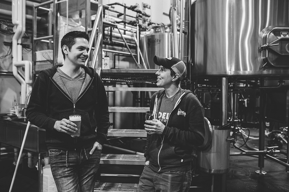 Two people drinking in front of industrial beer brewing equipment