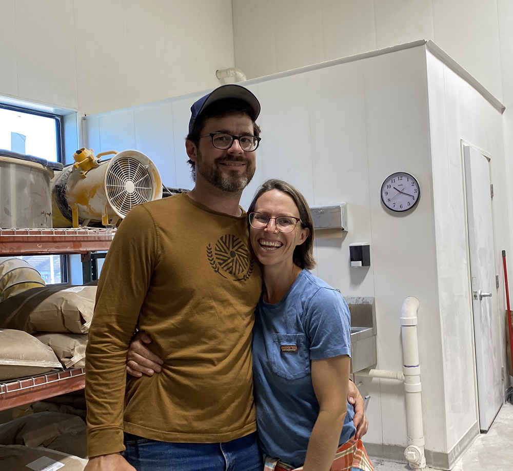 A couple with their arms around each other's waists in an industrial kitchen or storage area