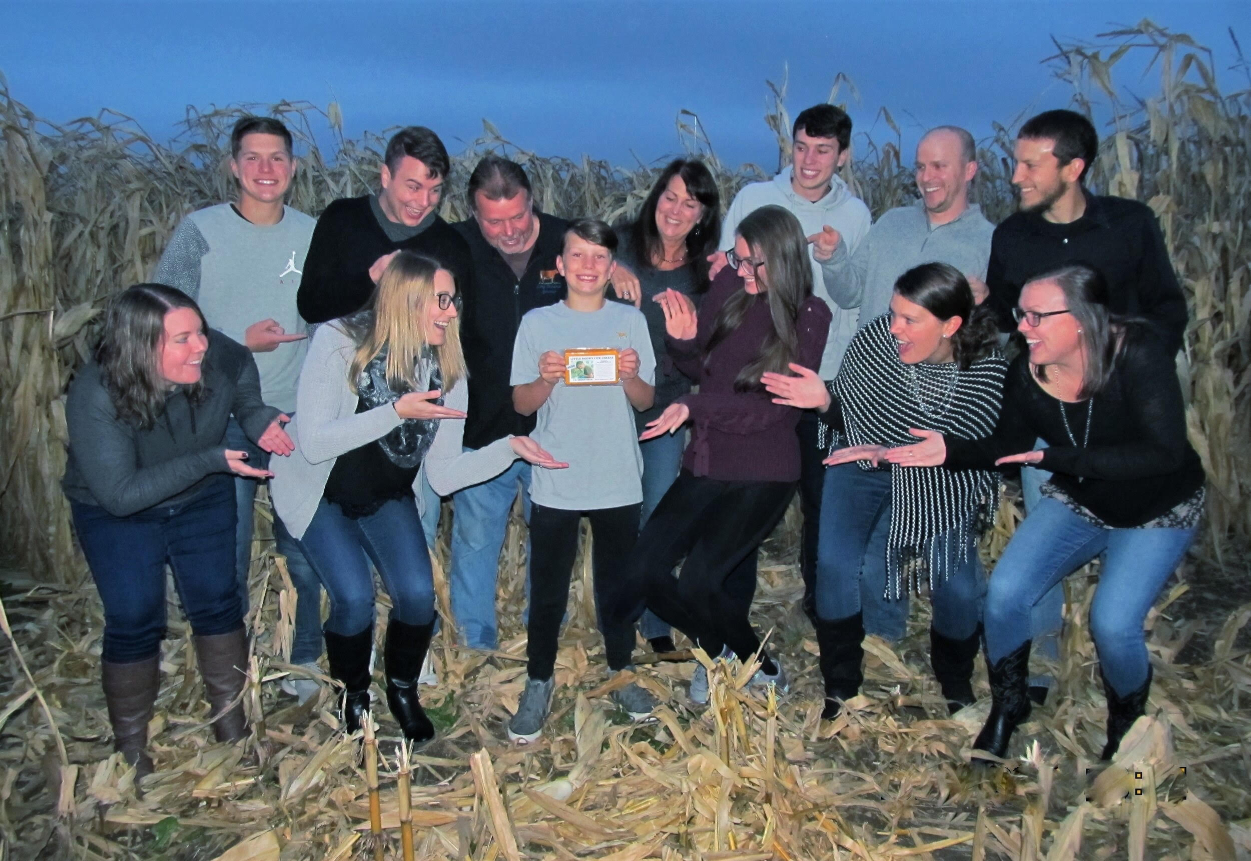 A group of people at dusk in a corn field pointing at a boy in the center holding a brick of cheese