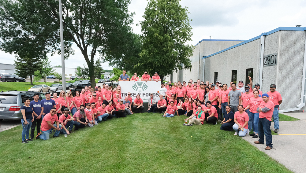 A large group of people posing around the Tribe 9 Foods sign outside the Monona Production Facility 