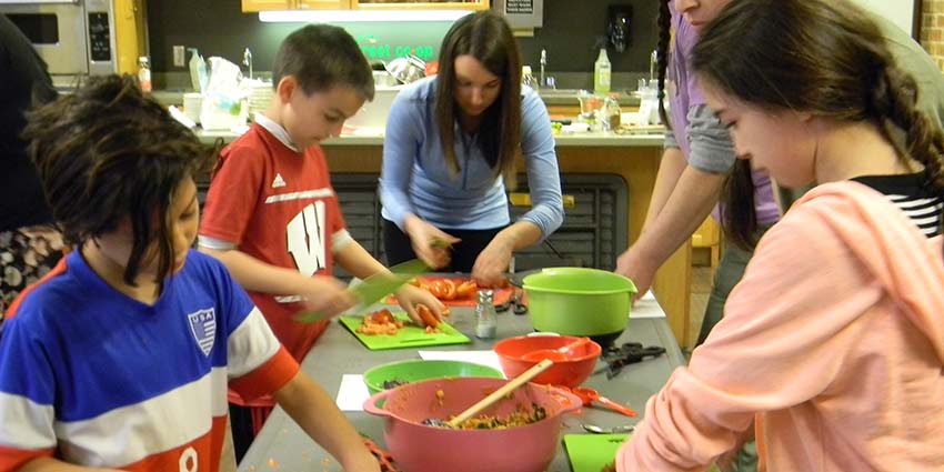 An instructor and kids cooking together at a long table