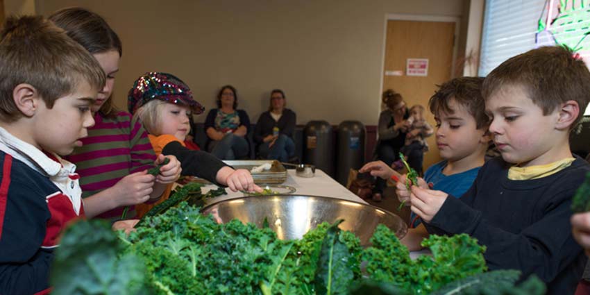 Small children working together at a long table to make something with kale