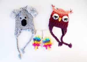 A knit hat with the appearance of a koala bear, a pair of white mittens with a rainbow design, and a knit hat in the shape of an owl