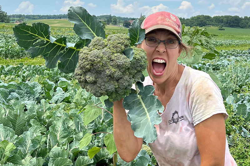 Cassie Noltnerwyss of Crossroads Community Farm holding a head of broccoli up to her head making an amazed face