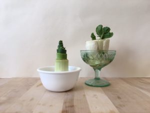 A white bowl holding a regrowing leek next to a stemmed glass holding regrowing bok choy