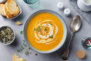 Pumpkin and carrot soup with cream, crusty bread, and toppings on grey stone background. Top view.