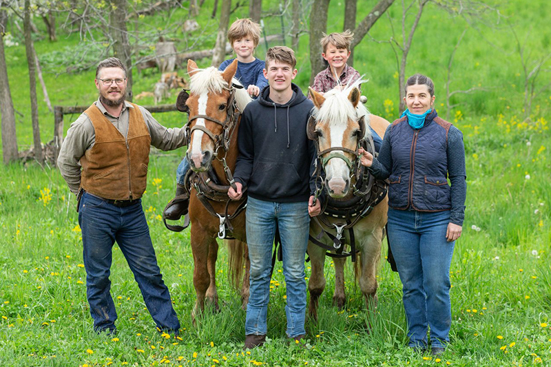 Robert and Summer Schultz and family of New Traditions Homestead posing in a field with two kids on ponies