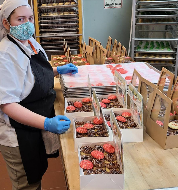 A person wearing gloves, an apron, a mask, and a hair net is packing up boxes of pastries