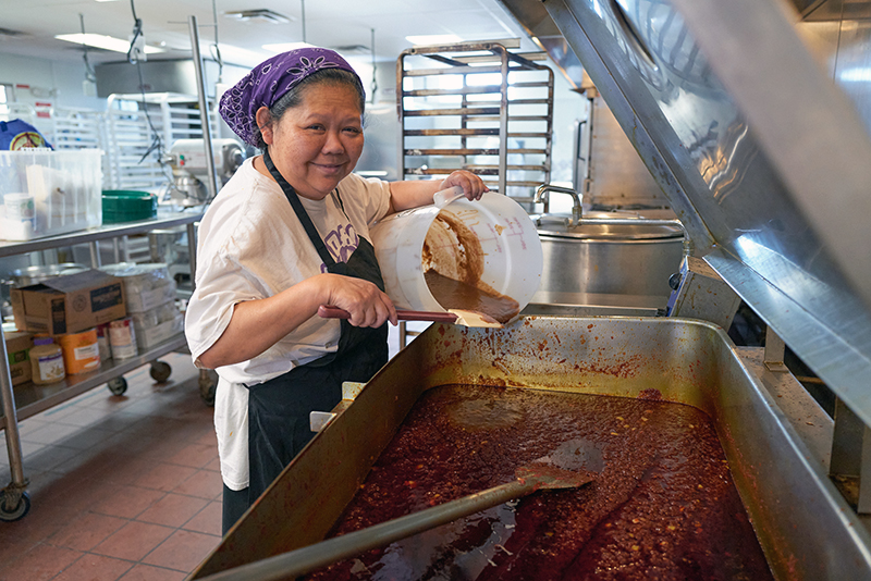 A woman working in an industrial kitchen pouring something into a vat