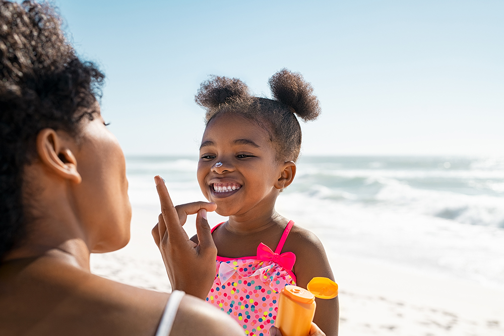 Young mother applying protective sunscreen on daughter nose at beach with copy space. Black woman hand putting sun lotion on female child face. African american cute little girl with sunblock cream at seaside.