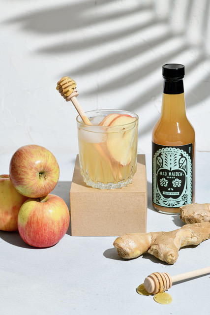 Product photo of a Mad Maiden shrub bottle next to a tumblr of a prepared drink and the ingredients used in the bottle